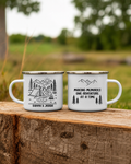 Two white enamel camping mugs with black printed graphics and text, displayed side-by-side on a rustic wooden log with a blurred outdoor background.