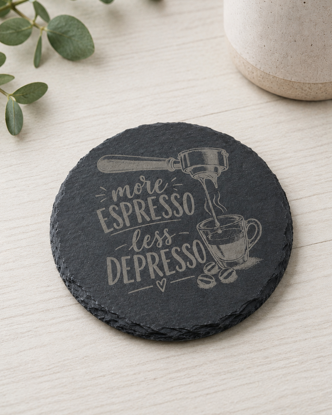 A round black slate coaster with a white engraved design featuring coffee illustrations and the phrase 'more ESpresso less DEPRESSO', shown on a light wood tabletop with decorative greenery and a ceramic mug in the background.