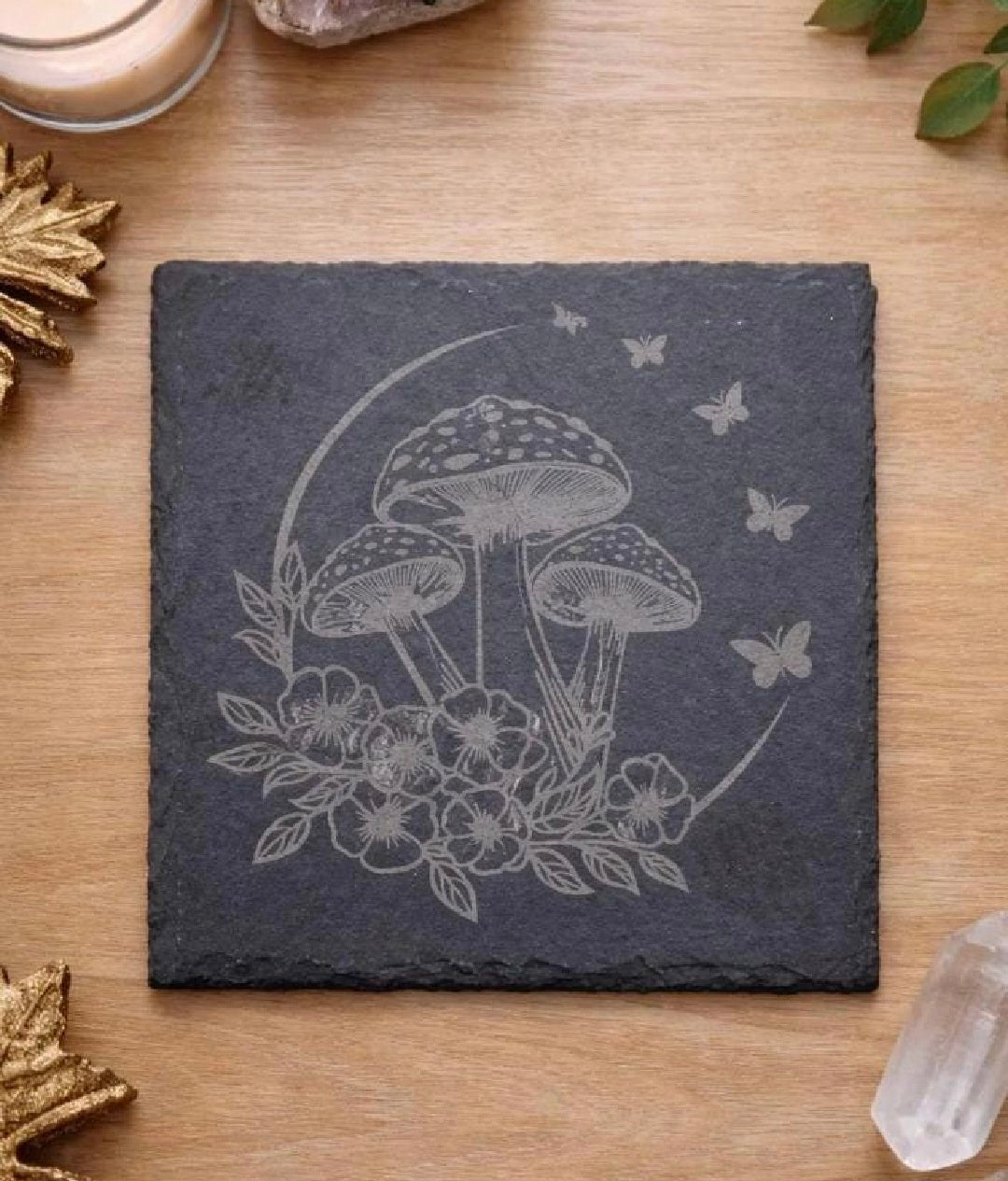 A square dark slate coaster with an engraved design of three mushrooms, flowers, leaves, and butterflies, displayed on a light wood tabletop with decorative props.