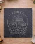 A square dark slate coaster featuring a light gray engraved design of three mushrooms, flowers, and a moon phase, displayed on a light wood tabletop with decorative crystals and leaves.