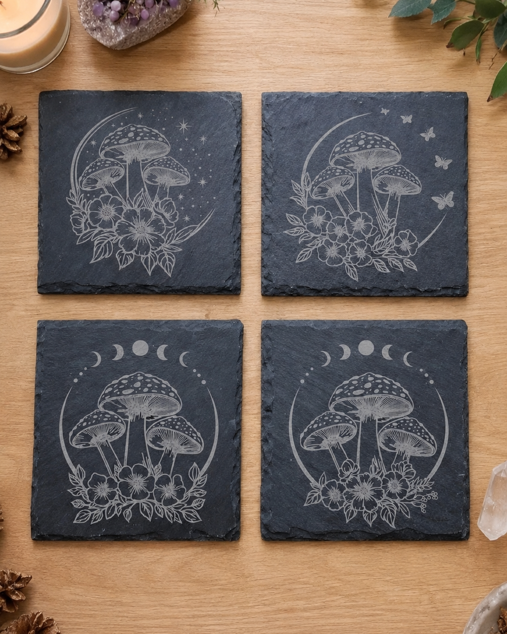 Four square dark slate coasters arranged in a 2x2 grid on a light wood surface, each printed with a white mushroom, moon phases, and floral design. A candle, crystals, and greenery are styled around the coasters.