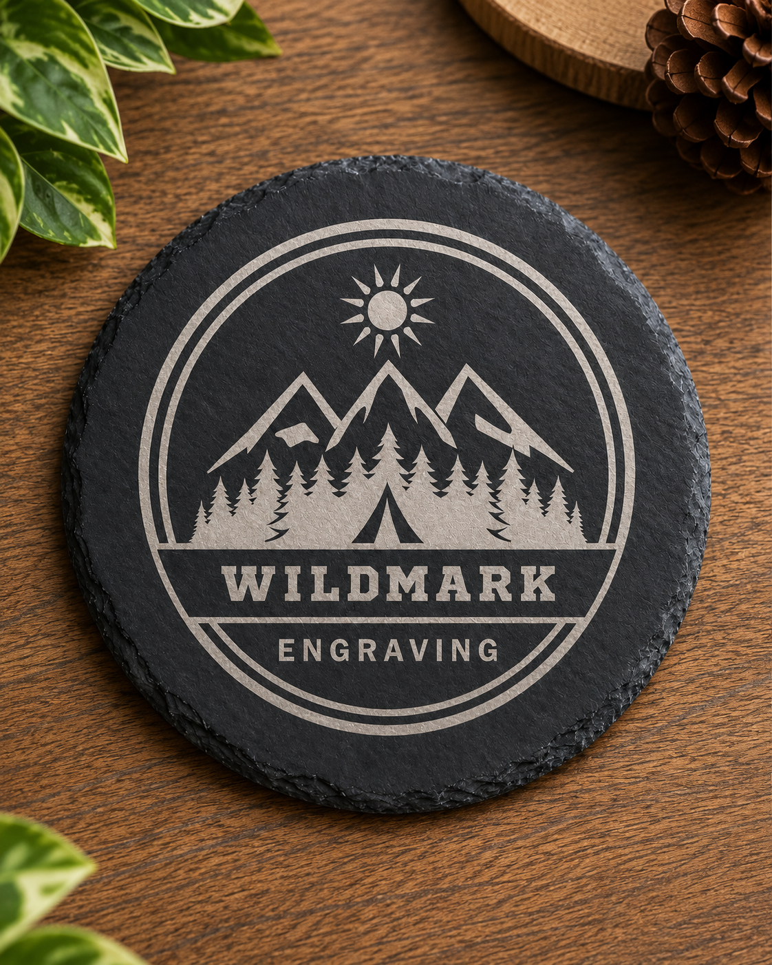A round black slate coaster with a white engraved mountain, sun, trees, and the words 'WILDMARK ENGRAVING,' placed on a wooden surface with leaves and a pinecone nearby.