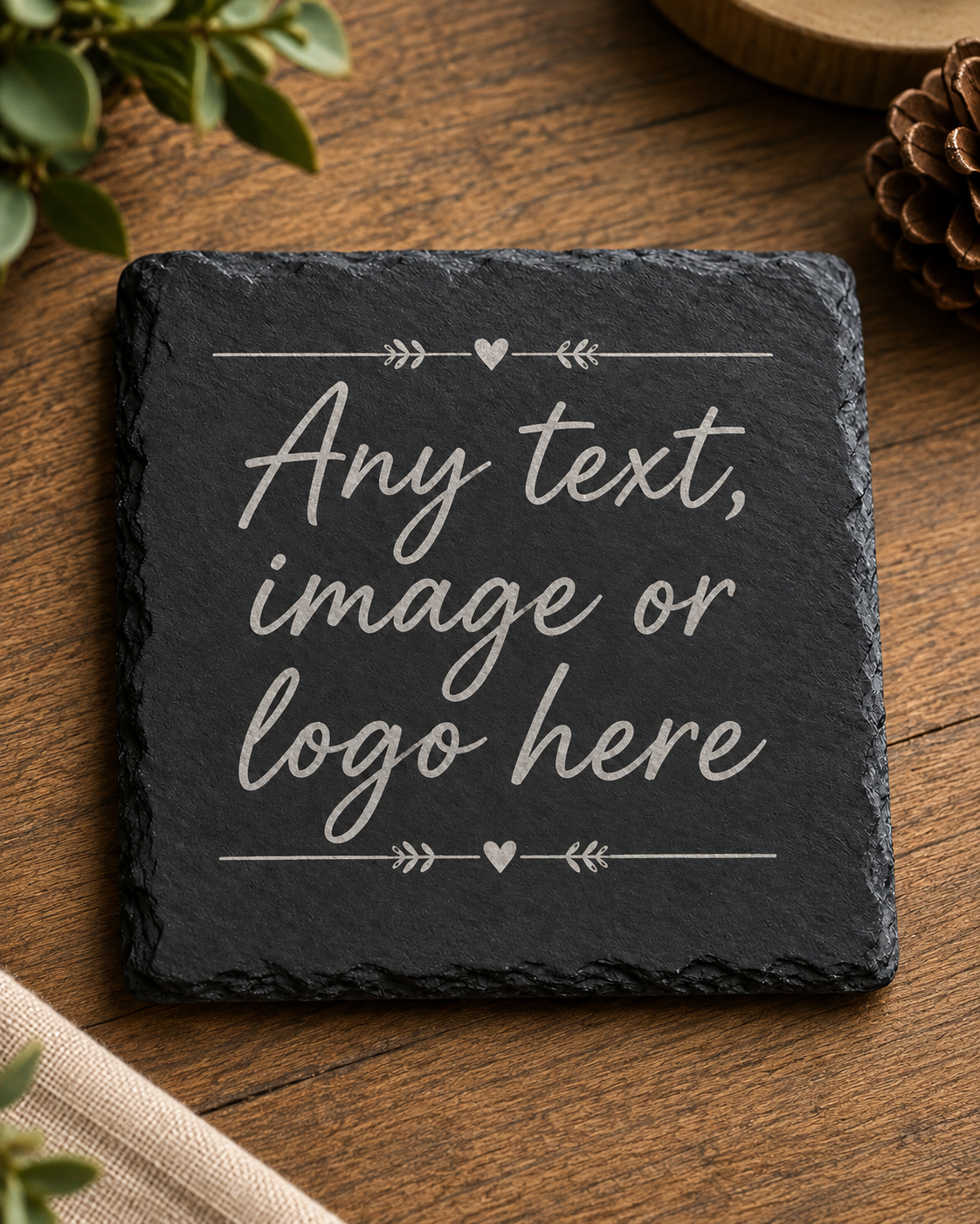 A square dark slate coaster with engraved white text reading 'Any text, image or logo here,' shown on a wooden tabletop with small decorative plants and a pinecone around it.