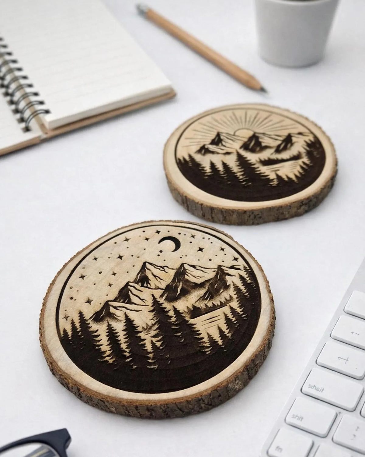Two round wood coasters with laser-engraved mountain and starry night designs, displayed on a white desk with a notebook, pencil, keyboard, and small plant in the background.