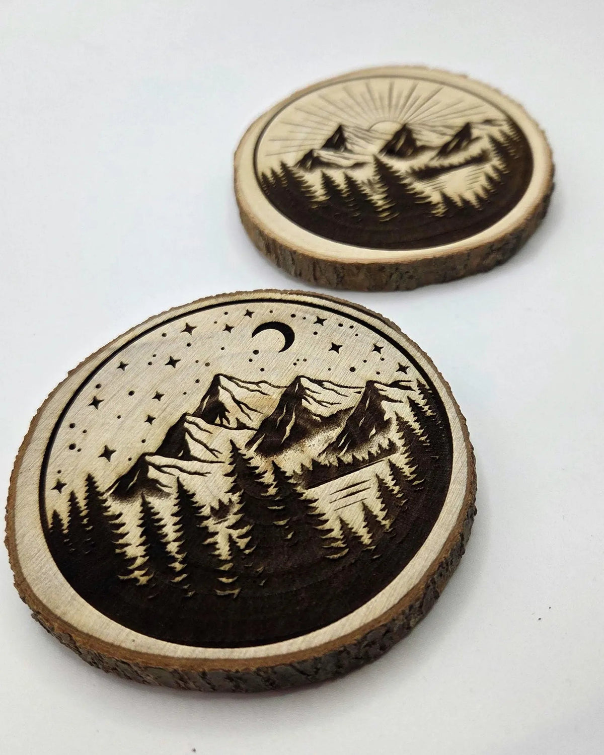 Two round wooden coasters with laser-engraved mountain and forest designs, displayed on a clean white background.