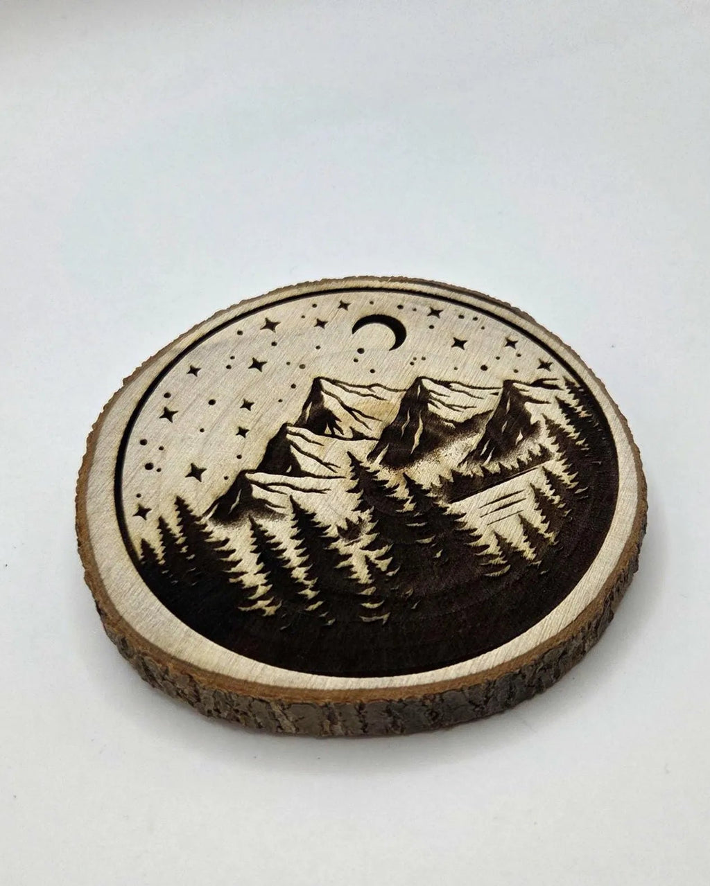 A round wooden coaster featuring a laser-engraved mountain and forest landscape under a crescent moon and stars, shown on a plain light background.