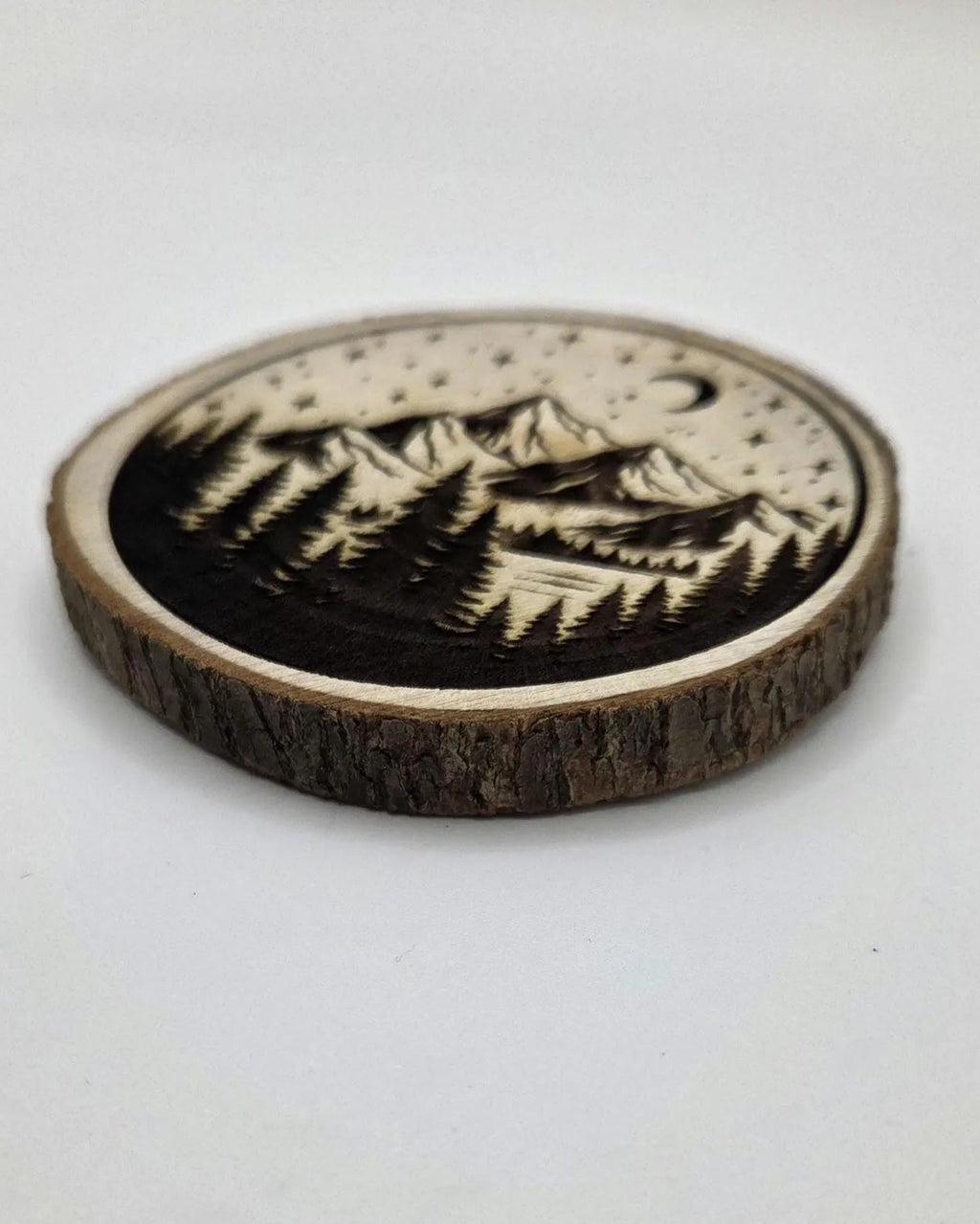 A round wooden coaster featuring a laser-burned mountain and pine forest landscape design with a crescent moon and stars, set against a plain light gray background.