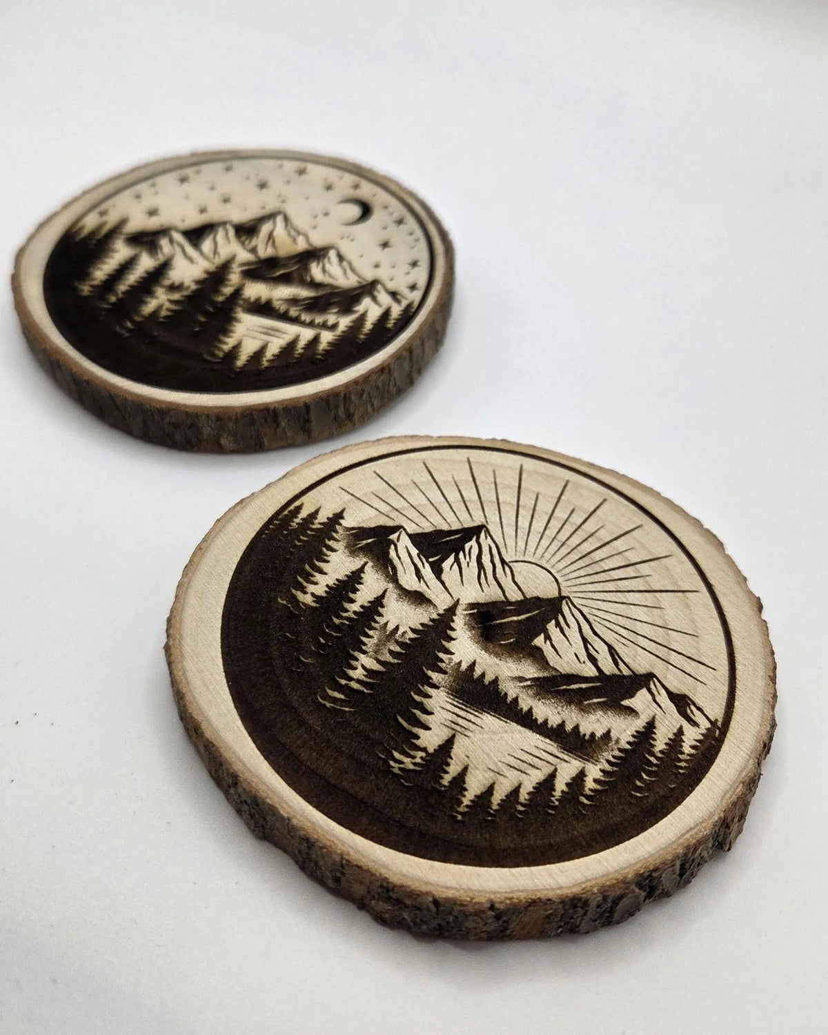 Two round natural wood coasters with engraved mountain and forest landscape designs, displayed on a plain white background.