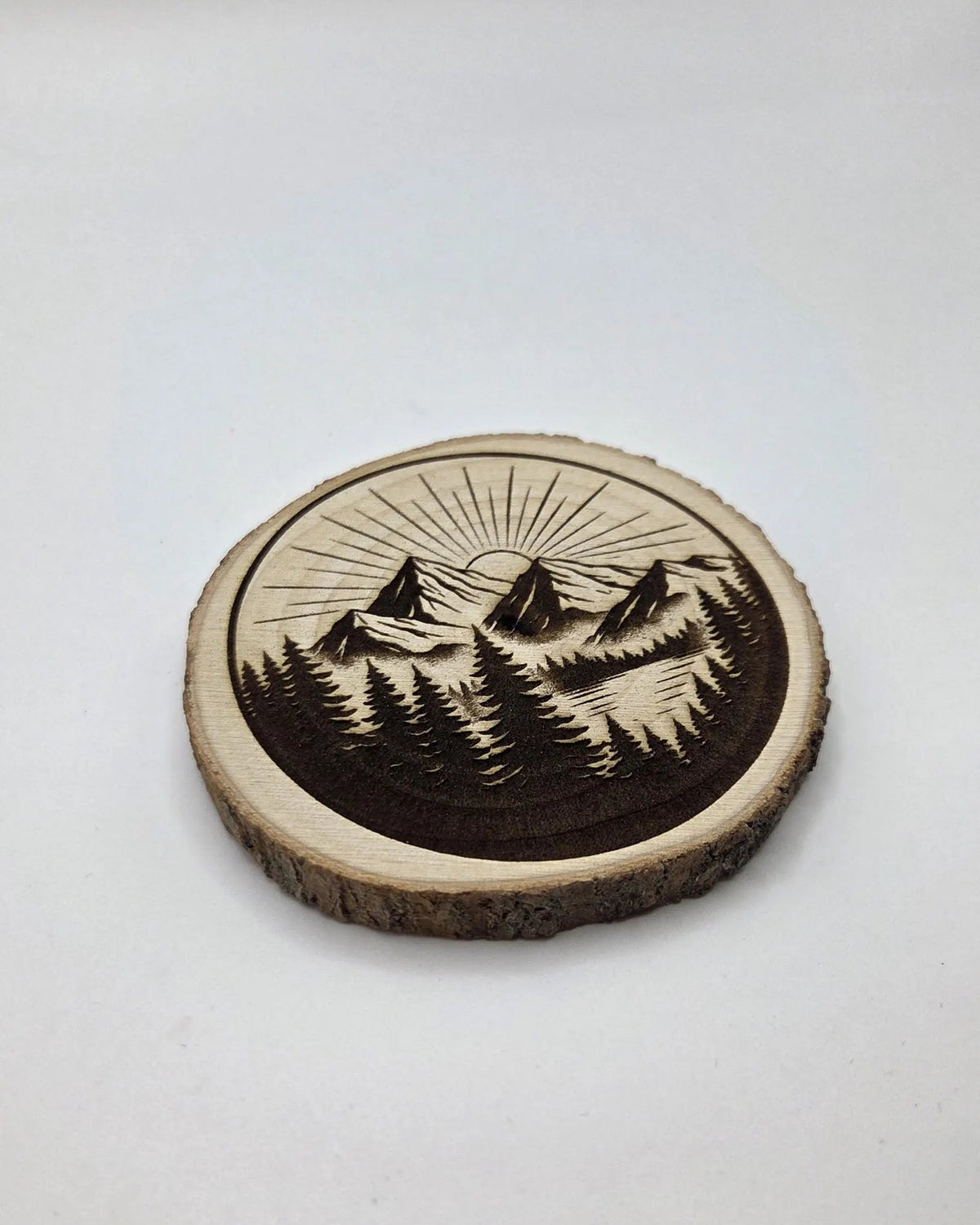 A round wooden coaster featuring a laser-engraved mountain and pine forest design, displayed on a neutral light gray background.